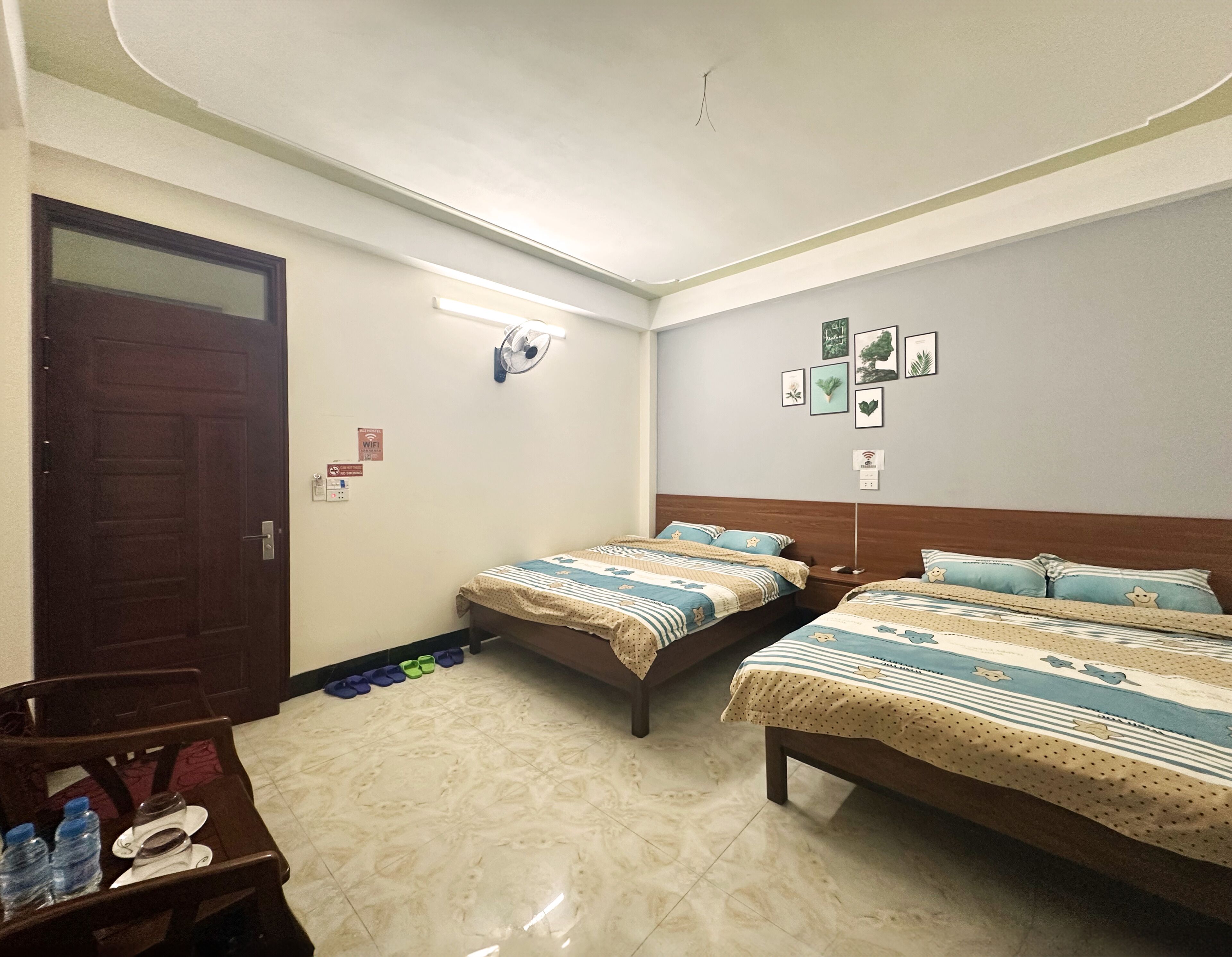 A hotel room with two beds, a wooden door, a fan, and a wall with framed pictures.