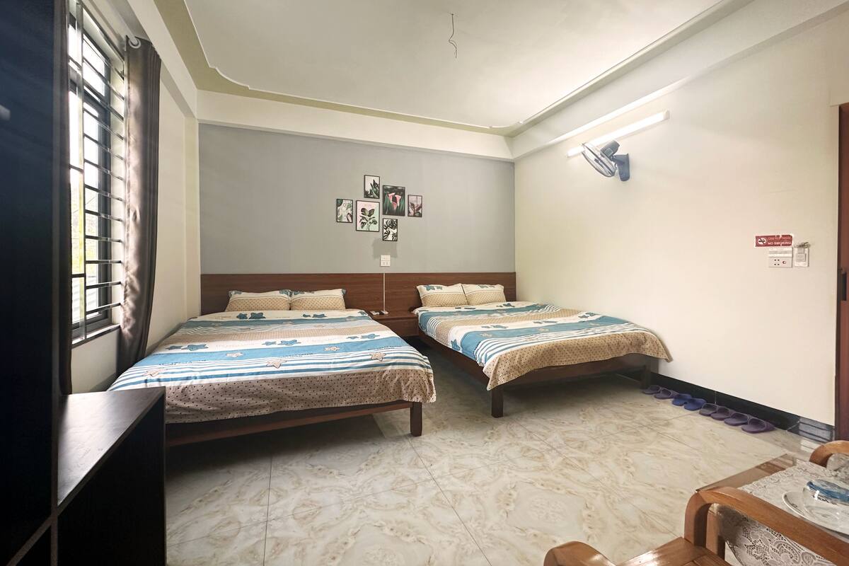 A hotel room with two beds, a small table, a chair, a window with curtains, a wall-mounted fan, and a ceiling light.