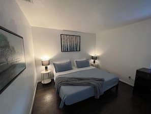 A bedroom with a bed, two bedside lamps, a painting on the wall, and a small table.