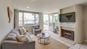 A living room with a sofa, armchair, glass coffee table, fireplace, and a TV mounted on the wall. - Oceanfront Paradise - AC, Private Patio, Gas Grill, Private Washer/Dryer! (San Diego)