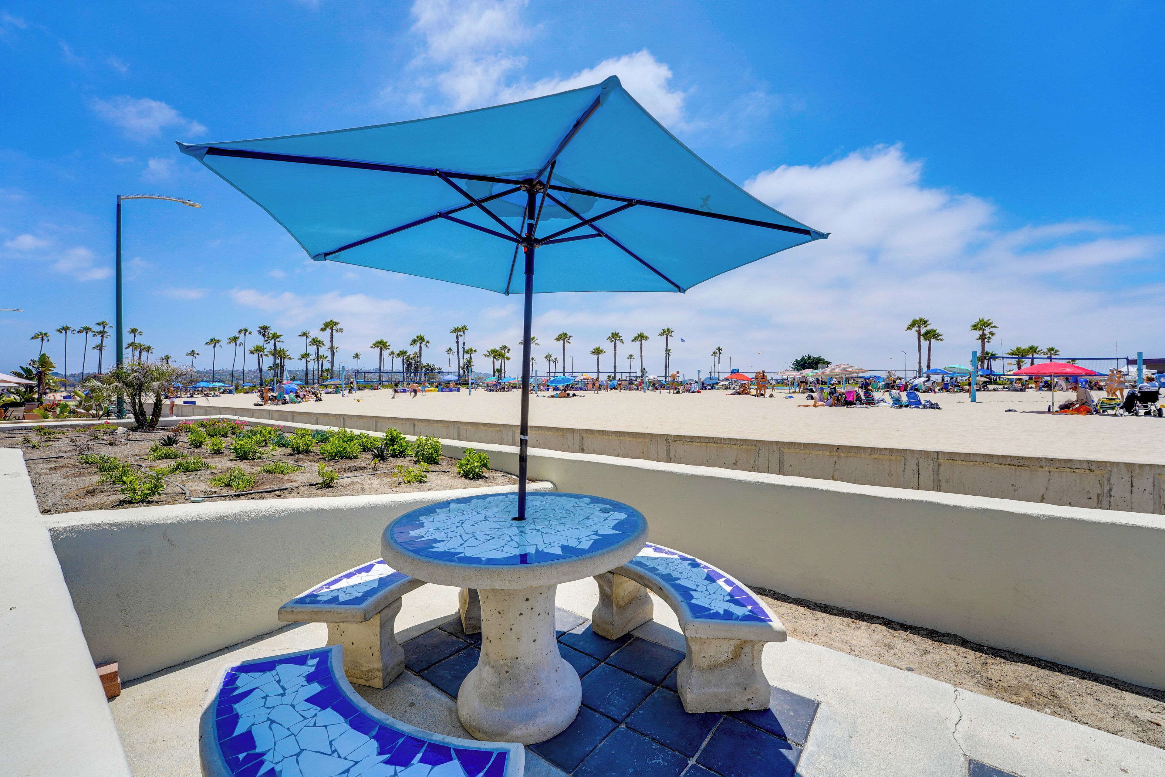 A beachside patio with a mosaic table and a blue umbrella, surrounded by palm trees and beach chairs.