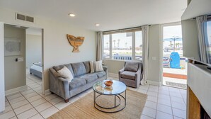 A modern living room with a grey sofa, a round coffee table, and a view of the beach through large windows. - Oceanfront Paradise - AC, Private Patio, Gas Grill, Private Washer/Dryer! (San Diego)