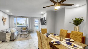 A modern living room with a sofa, a dining table set for four, and a TV mounted on the wall. - Oceanfront Paradise - AC, Private Patio, Gas Grill, Private Washer/Dryer! (San Diego)