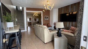 Un salon moderne avec un canapé, une table basse et un téléviseur fixé au mur. Un escalier mène à un autre niveau, et il y a un coin repas avec des chaises et une table.