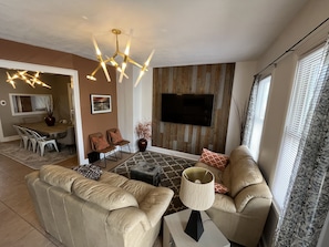 Una sala de estar moderna con un sofá, una mesa de centro y un televisor instalado en una pared de madera. Se ve un comedor al fondo.