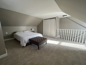 Un dormitorio con cama, reposapiés, mesita de noche, lámpara y baranda blanca.