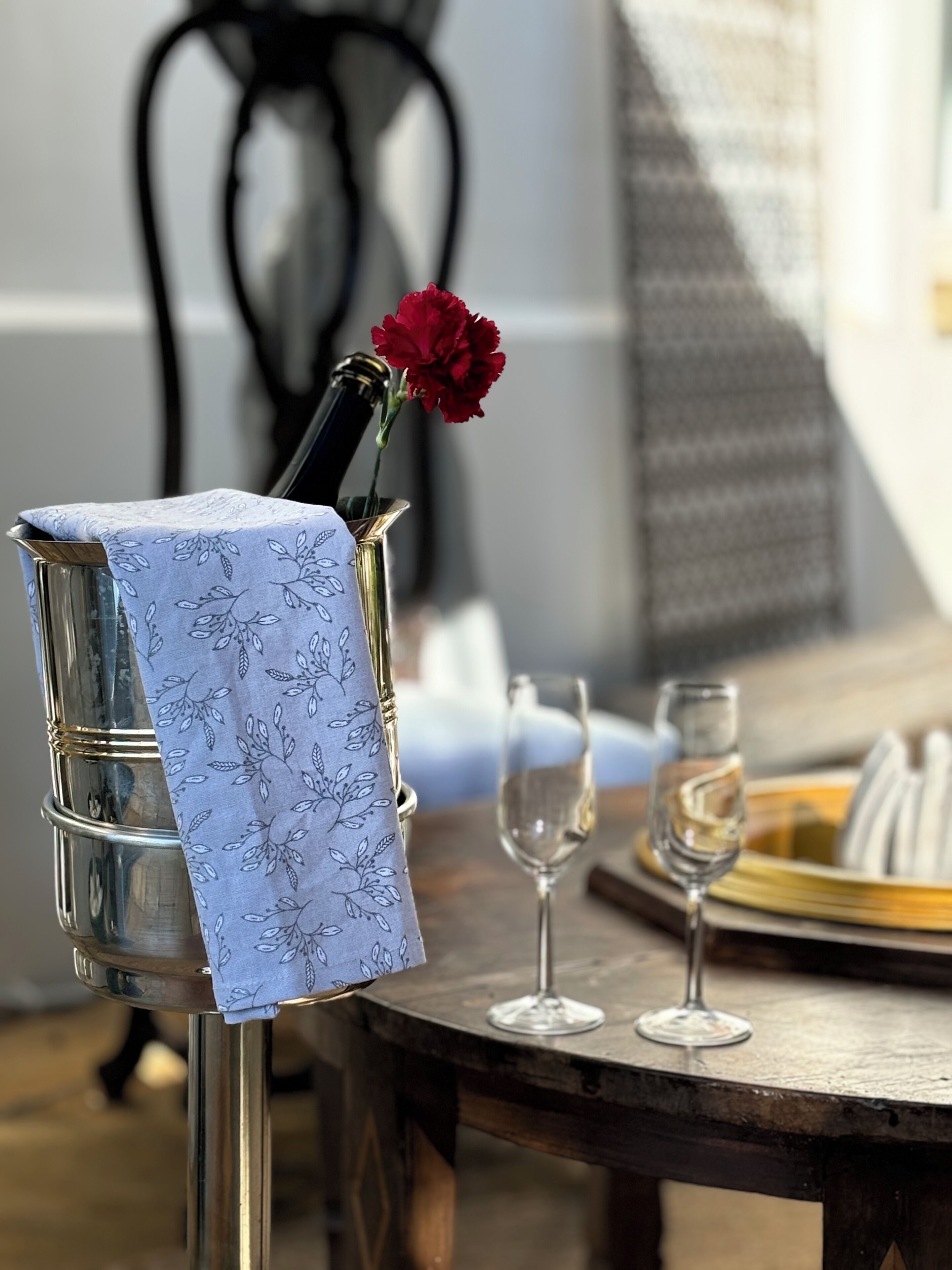 Un vase en verre contenant une fleur rouge, posé sur un support en métal, un verre à vin et une assiette avec de la nourriture, le tout sur une table en bois.