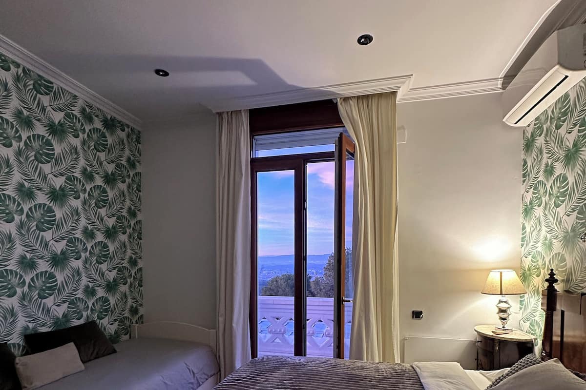 A hotel room with two beds, a view through an open window, and a nightstand with a lamp.