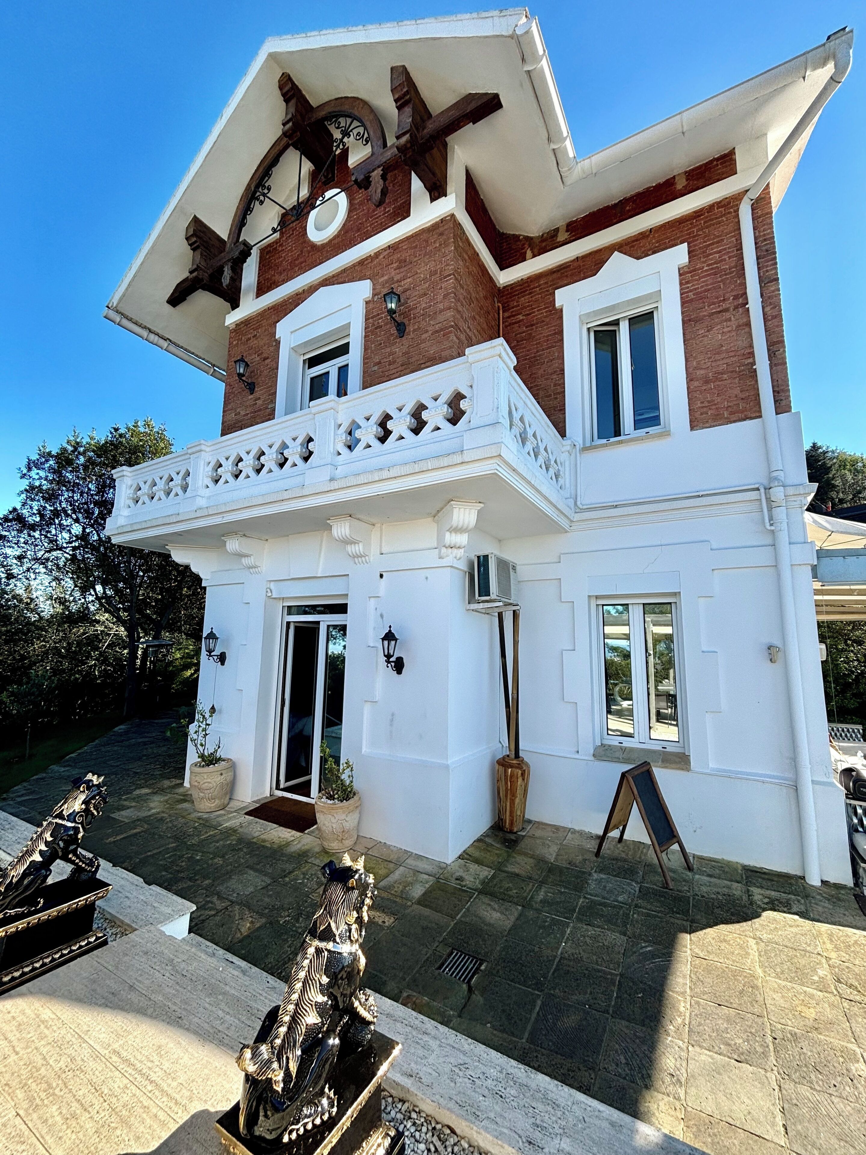 Un edificio de dos pisos con fachada blanca y detalles de ladrillo marrón, que presenta un balcón con barandas decorativas y una estructura de madera sobre la entrada.