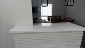 A kitchen with a white brick accent wall, a white countertop, and a window with blinds.