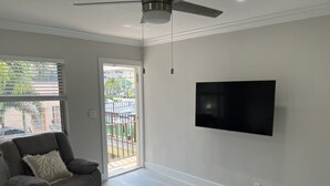 A spacious living room with a grey recliner, a flat-screen TV mounted on the wall, and a sliding glass door leading to an outdoor balcony.