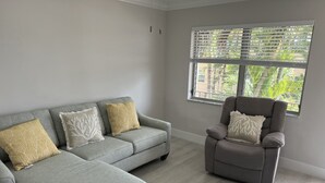 A living room with a grey sofa and armchair, two patterned cushions, and a window with blinds.