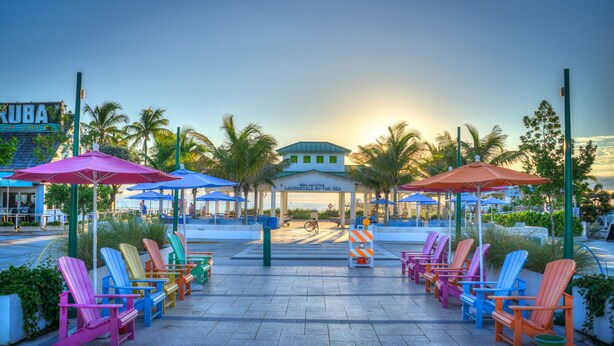A beachfront area with colorful Adirondack chairs and umbrellas, a sign for 'LAUDERDALE-BY-THE-SEA', and palm trees.