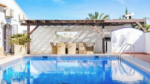 A swimming pool with a tiled edge, a pergola with seating, and a white wall with a window. - Village La Gomera (Jávea)