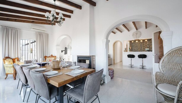 A dining area with a wooden table set for six, grey upholstered chairs, and a chandelier. An archway leads to a kitchen area with a clock and bar stools. - Village La Gomera (Jávea)