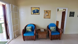 Una habitación con dos sillones de mimbre con cojines azules, una mesita con una revista y una puerta que da a otra habitación.