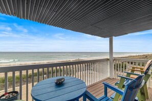 Een houten terras met een blauwe tafel en groene en blauwe stoelen, met uitzicht op een strand, de oceaan en een heldere lucht.