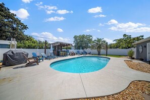 Un patio trasero con piscina, sillas de descanso, zona de barbacoa y parrilla cubierta.