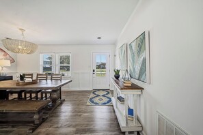 A dining area with a wooden table and chairs, a chandelier, and a view through a door to the outside. - The Oasis - Heated POOL - Tiki Bar - Beach Life! (Carolina Beach)