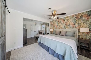 Ein Schlafzimmer mit einem Bett, einem Deckenventilator, einem Nachttisch, einer Lampe, einem Sessel und einer Tür zu einem anderen Raum.