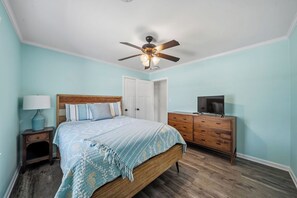Un dormitorio con cama, cómoda de madera, televisor, mesita de noche, lámpara y ventilador de techo.