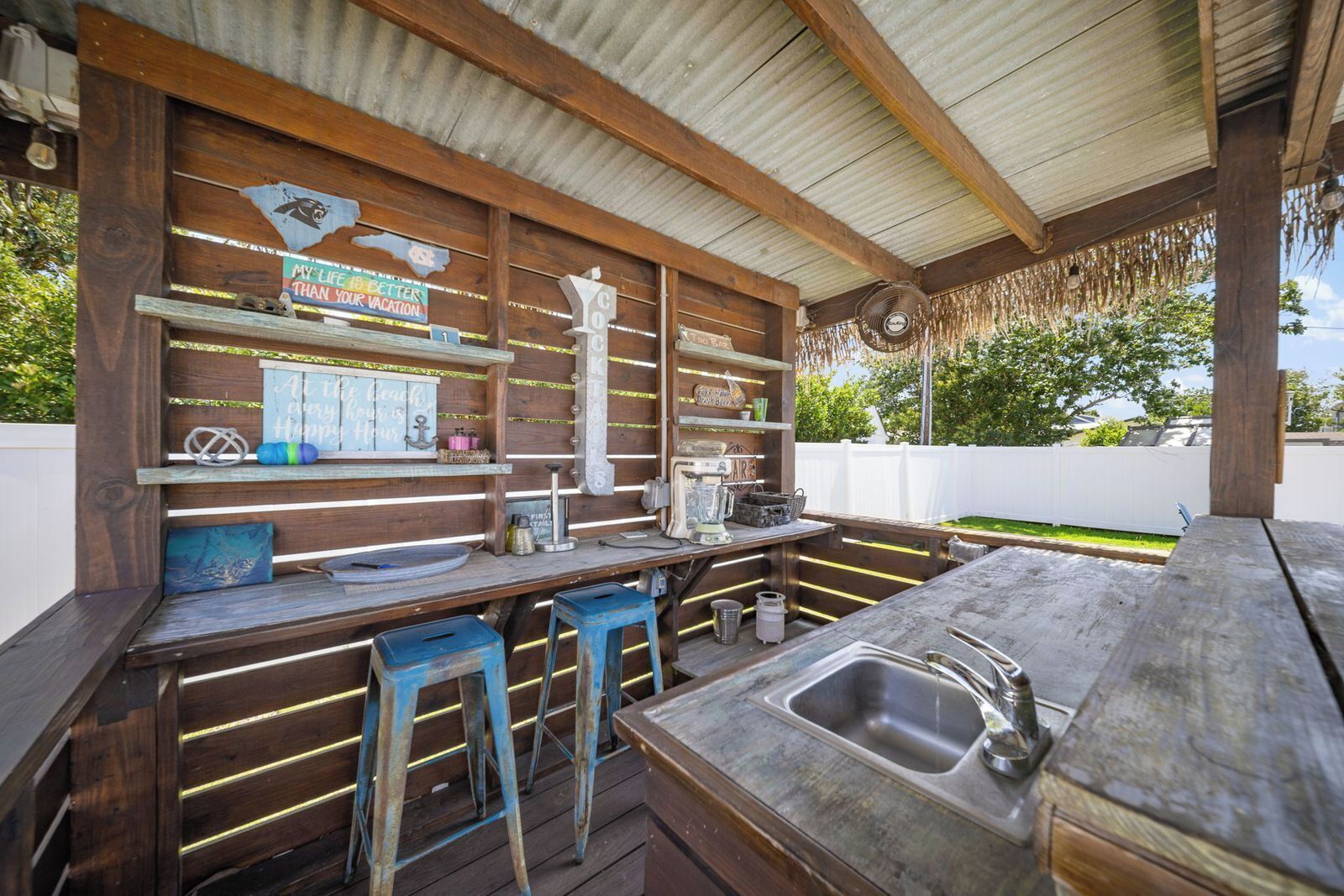 A rustic outdoor kitchen with wooden beams, a sink, stools, and various kitchen appliances.