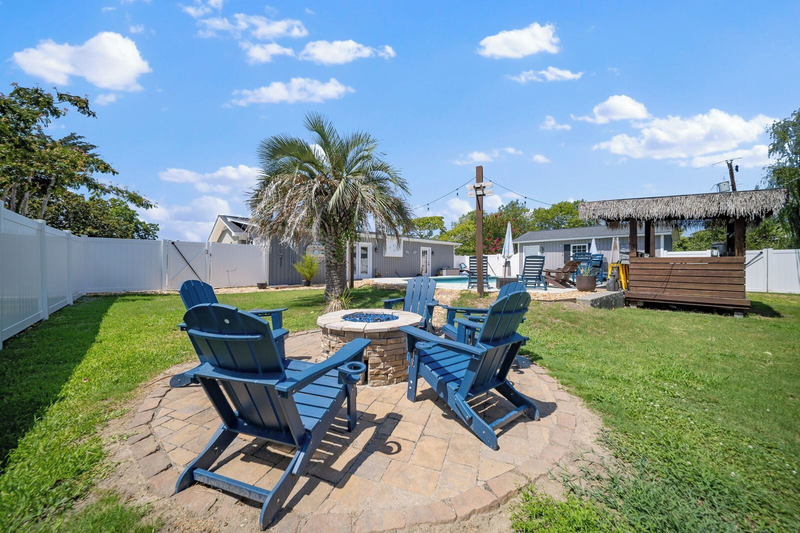 A backyard with a stone patio, two Adirondack chairs, a small fire pit, and a pool area in the background.