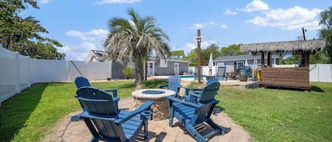 A backyard with a stone patio, two Adirondack chairs, a small fire pit, and a pool area in the background.