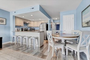 Una cocina con armarios blancos, una isla central con taburetes, una mesa de comedor con sillas y un refrigerador.
