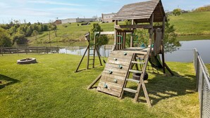 A wooden play structure with a slide and climbing ladder, surrounded by a fenced grassy area and a body of water. - 4 Bed, 2.5 Bath, 3 KING BEDS on a Private Lake with Dock! Family Friendly (Crittenden)