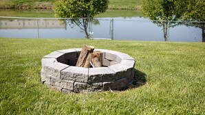 A circular stone fire pit with logs in a grassy area near a body of water and a fence. - 4 Bed, 2.5 Bath, 3 KING BEDS on a Private Lake with Dock! Family Friendly (Crittenden)
