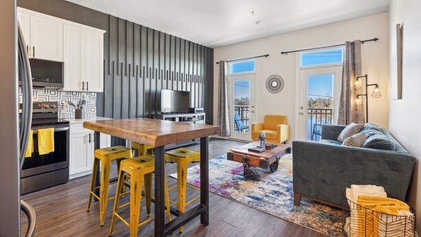 A modern kitchen with white cabinets, a wooden table, and yellow stools. There is a grey sofa and a coffee table in the living area.
