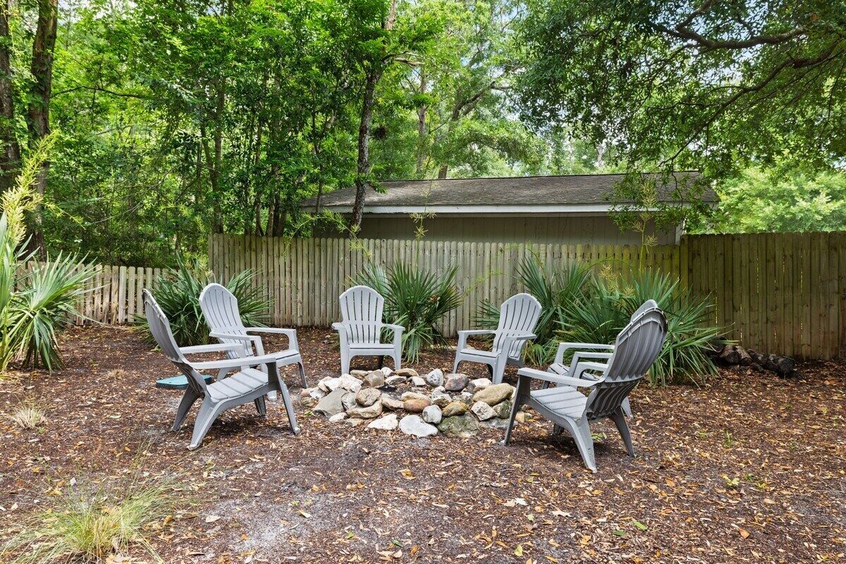 A backyard with Adirondack chairs arranged in a circle around a stone fire pit, surrounded by a wooden fence and lush greenery.