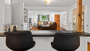 A modern living room with a sofa, coffee table, and a dining area with chairs. There is a bookshelf and a window with curtains. - Rising Sun Retreat - 4 Miles to Beach (Wilmington)