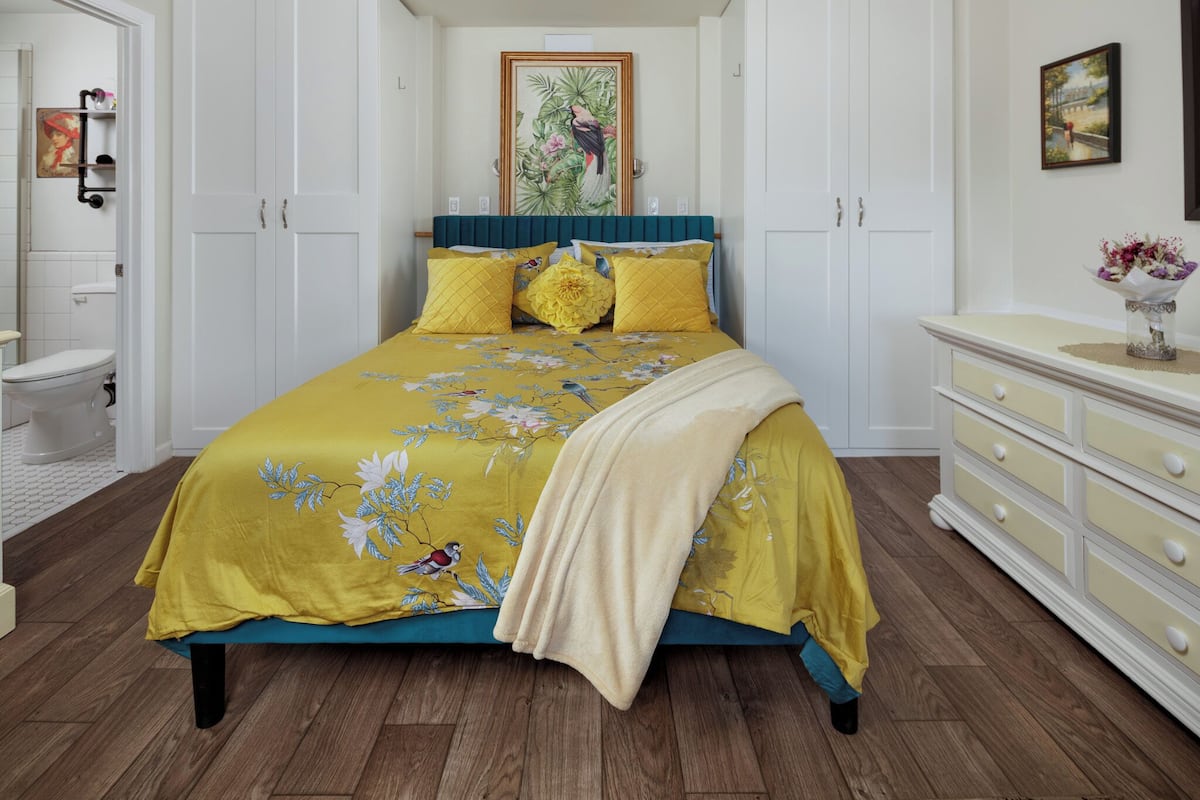 A bedroom with a bed, a dresser, a painting on the wall, and a bathroom visible in the background.