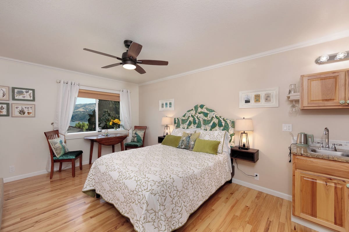 A bedroom with a bed, a desk, a chair, a nightstand, a lamp, a ceiling fan, and a view of the outdoors.