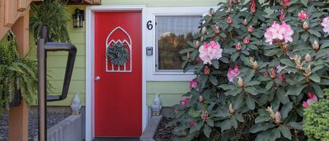 Una puerta roja con una corona, el número '6' en la parte superior y un arbusto de flores rosas al frente.
