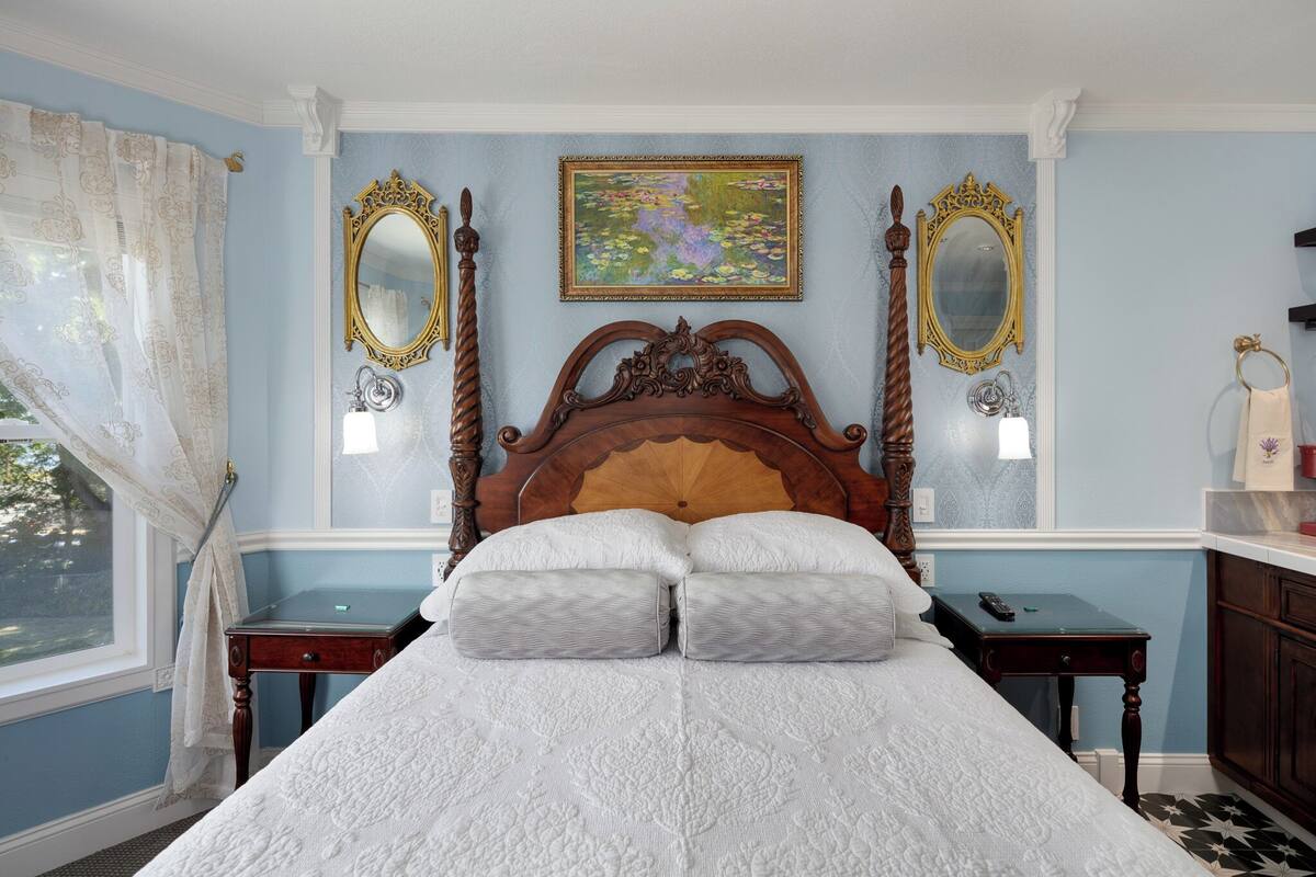 A bedroom with a large bed, two bedside tables, a window with curtains, and two wall-mounted mirrors.