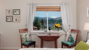 Une pièce avec vue sur les montagnes et un lac, deux fauteuils agrémentés de coussins décoratifs, une petite table avec une bouteille et des verres, et des reproductions botaniques encadrées accrochées au mur.