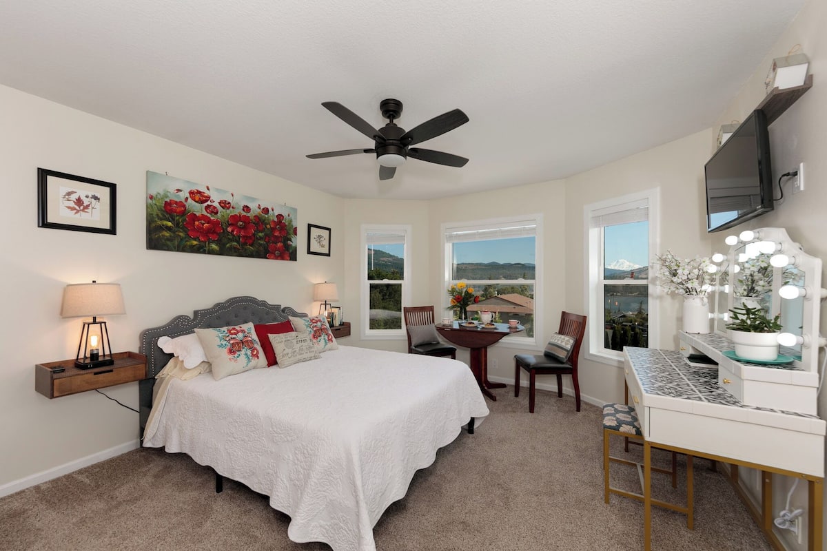 A bedroom with a bed, a desk with a vanity, a ceiling fan, and large windows.