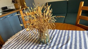 Ruokapöytä, jonka päällä on raidallinen pöytäliina, maljakko, jossa on kuivattuja vehnänkorvia, ja vihreällä sohvalla taustalla on sinivalkoraidallinen koristetyyny.