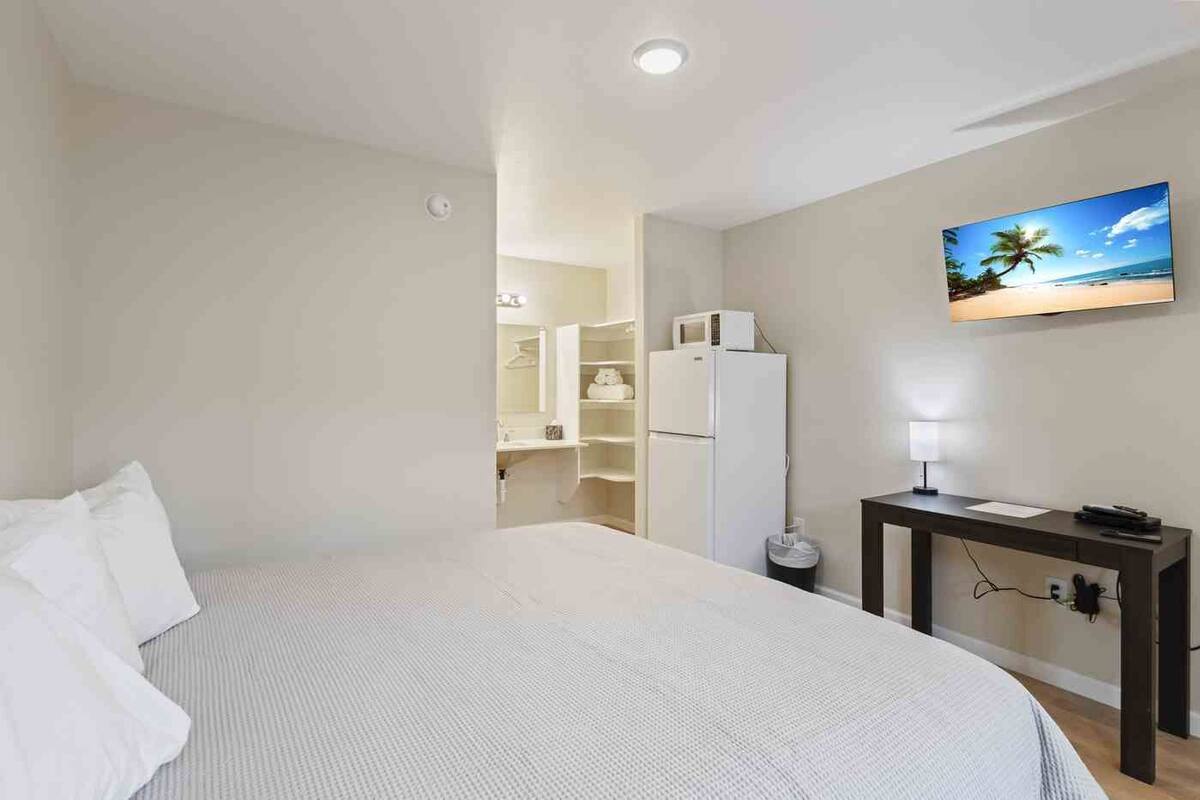 A bedroom with a bed, a desk with a lamp, a refrigerator, and a TV on the wall.