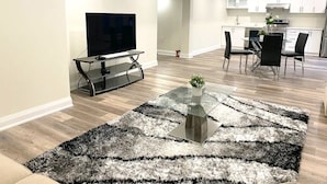 A modern living room with a flat-screen TV, a glass coffee table with a plant, and a dining area with a kitchen in the background. - Brand New Walk Out Bsmt Apt Near Beach  (Pickering)