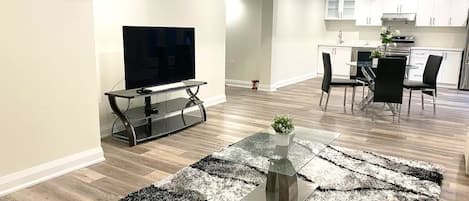 A modern living room with a flat-screen TV, a glass coffee table with a plant, and a dining area with a kitchen in the background.