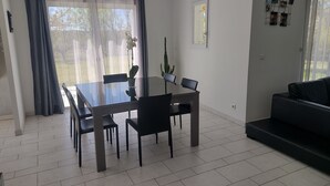 Un salon moderne avec un coin repas, comprenant une table à plateau en verre, des chaises noires, un canapé noir et une grande fenêtre ornée de rideaux légers.