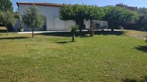 Un bâtiment blanc avec un toit en tuiles, entouré d’une zone herbeuse, d’arbres et d’un mur de pierre.