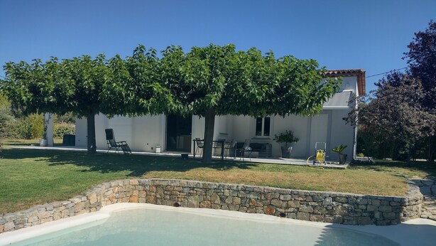 Une piscine bordée de pierres, une cour gazonnée et un bâtiment blanc agrémenté de mobilier d’extérieur.