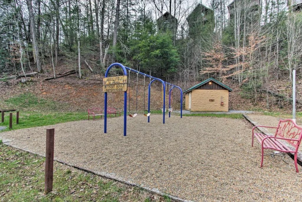 A playground with swings and a bench, surrounded by trees and a wooden structure.