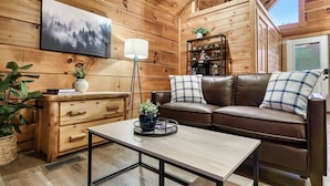 A cozy wooden cabin interior with a brown leather sofa, a wooden dresser, a coffee table, and a bookshelf. - 10 Min to Gatlinburg Strip Hot Tub King Bed Pool, 5 min to Rocky Top Sport World (Gatlinburg)