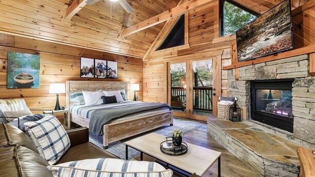 A cozy log cabin interior with a large bed, a fireplace, and a stone wall. - 10 Min to Gatlinburg Strip Hot Tub King Bed Pool, 5 min to Rocky Top Sport World (Gatlinburg)